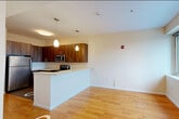 Located at 100 Newkirk Street in Jersey City, NJ, this efficiently designed single-floor unit offers a well-organized layout comprising a kitchen, hallway, and full bathroom. The kitchen is a standout feature, outfitted with rich dark walnut-toned cabinetry — both upper and lower — paired with light quartz countertops for a modern contrast. Stainless steel appliances, including an electric range, built-in microwave, dishwasher, and refrigerator, provide a fully equipped cooking space. Pendant lighting adds a contemporary touch above the kitchen area. The main living space features warm hardwood flooring that extends through the hallway, creating a cohesive flow throughout the unit. A large window frames an expansive cityscape view, filling the room with natural light and offering a scenic backdrop. The bathroom is cleanly finished with white cabinetry, a grey countertop vanity, and a full soaking tub and shower combination accented by distinctive seafoam green mosaic tile that lines the walls, adding a pop of color. Neutral white walls and tile flooring complete the bathroom's clean aesthetic. The entry hallway, finished with the same hardwood flooring, connects all areas of the unit seamlessly, making efficient use of the available space.
