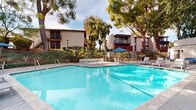 This outdoor pool area serves as a central amenity within a residential community, offering a well-maintained and inviting environment for recreation and relaxation. The pool itself is notably spacious, featuring clear turquoise water and a classic geometric shape with entry steps and safety rails for easy access. Depth markers are visible along the pool's edge, and a life ring station adds a practical safety element.

The surrounding deck is finished in smooth concrete and furnished with modern slatted lounge chairs and small side tables, providing comfortable spots for sunbathing. A blue striped patio umbrella shades a bistro-style table and chair set, offering a shaded seating area ideal for casual gatherings. The entire pool area is enclosed by a black metal security fence with a keyed-access gate, ensuring privacy and safety.

Mature trees—including large oaks and pines—frame the space, casting dappled shade and contributing to a park-like atmosphere. Lush hedging and ornamental plantings line the perimeter, adding greenery and visual softness. The surrounding multi-story buildings feature exterior staircases and private balconies, visible from the pool deck. Abundant natural light fills the space throughout the day, creating a bright and open outdoor ambiance well-suited for year-round enjoyment in a mild climate.