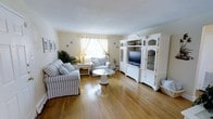 This charming single-floor space offers a warm and inviting atmosphere highlighted by rich hardwood flooring that flows throughout. The living area serves as the heart of the home, thoughtfully arranged with blue-and-white striped upholstered seating that lends a classic coastal character to the room. A round glass-topped coffee table anchors the seating arrangement, adding a touch of elegance without overwhelming the space. Natural light filters in through a window dressed with soft yellow curtains, brightening the neutral-toned walls and creating a cheerful, open ambiance. A large white entertainment unit with glass-front cabinetry stands as a prominent architectural feature along one wall, offering both display and storage space while contributing to the room's clean, traditional aesthetic. The unit's arched detailing and ornate molding add a refined, timeless quality to the interior. Lush green houseplants are thoughtfully placed to bring life and vibrancy to the corners of the room. The overall design is cohesive and serene, combining classic furnishings with light, neutral tones to create a comfortable and well-appointed living environment that feels both functional and welcoming.