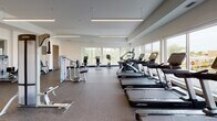 Located at 12501 Ridgedale Dr in Minnetonka, Minnesota, this spacious fitness center offers a well-appointed, single-floor layout designed to support a full range of workout routines. The facility is bright and open, benefiting from large windows along multiple walls that flood the space with natural light and provide views of the surrounding outdoor environment.

The cardio zone is equipped with a long row of Life Fitness treadmills, stationary bikes, and elliptical machines, all positioned along the windowed perimeter to enhance the exercise experience. A dedicated free weights area features adjustable benches, a squat rack, dumbbells, and a rowing machine, accompanied by a wall-mounted flat-screen television and mirrored walls that visually expand the space.

The strength training section includes a variety of cable and selectorized machines arranged throughout the floor, offering targeted training options for all major muscle groups. Mirrors line portions of the walls to support proper form and technique. The durable rubber flooring throughout provides both safety and comfort underfoot.

Overhead, linear pendant lighting fixtures complement the natural light, ensuring the entire facility is consistently and evenly illuminated. The clean white walls and modern finishes contribute to an organized, professional atmosphere conducive to focused training.