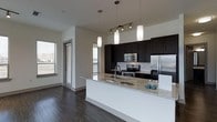 This single-floor space showcases a thoughtfully designed open-concept layout centered around a sleek, modern kitchen. The kitchen is anchored by an expansive island topped with light granite countertops, offering generous prep and seating space. Dark espresso-toned cabinetry provides a bold contrast against the white subway tile backsplash and crisp white walls, creating a striking visual balance. Stainless steel appliances, including a refrigerator, range, and built-in microwave, lend a polished, professional-grade aesthetic. Overhead, a combination of pendant lights and track lighting illuminates the space effectively, adding both function and style. Rich, dark hardwood flooring runs throughout, grounding the space with warmth and continuity. Multiple windows allow natural light to filter in from several angles, brightening the interior and contributing to an open, airy ambiance. The overall design blends contemporary style with practical functionality, making the kitchen the clear focal point of the space while maintaining a cohesive and inviting atmosphere throughout.