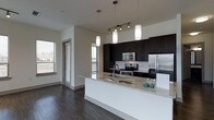 This single-floor space showcases a thoughtfully designed open-concept layout centered around a sleek, modern kitchen. The kitchen is anchored by an expansive island topped with light granite countertops, offering generous prep and seating space. Dark espresso-toned cabinetry provides a bold contrast against the white subway tile backsplash and crisp white walls, creating a striking visual balance. Stainless steel appliances, including a refrigerator, range, and built-in microwave, lend a polished, professional-grade aesthetic. Overhead, a combination of pendant lights and track lighting illuminates the space effectively, adding both function and style. Rich, dark hardwood flooring runs throughout, grounding the space with warmth and continuity. Multiple windows allow natural light to filter in from several angles, brightening the interior and contributing to an open, airy ambiance. The overall design blends contemporary style with practical functionality, making the kitchen the clear focal point of the space while maintaining a cohesive and inviting atmosphere throughout.