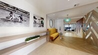 This single-floor hallway and lobby space offers a thoughtfully designed interior that blends contemporary aesthetics with functional flow. The layout transitions from a welcoming lobby area into an extended corridor, creating a sense of depth and movement throughout the space.

The lobby area immediately establishes a strong visual identity through its herringbone-patterned wood-look flooring, which anchors the space warmly. A striking teal pendant chandelier serves as a focal point overhead, while a vibrant mustard-yellow channeled sofa provides bold contrast against the crisp white walls. Built-in wood shelving integrated into the walls displays potted greenery, adding organic texture. Large-scale black-and-white photography and a gold-framed oval mirror contribute curated artistic character.

A geometric wood-panel divider with a colorful fiber art installation introduces an expressive, contemporary accent, while black-framed glass doors and floor-to-ceiling windows along the corridor allow natural light to filter through and offer views of an outdoor area. The extended hallway features light-toned porcelain tile flooring, recessed ceiling lighting, and clean white walls, maintaining a bright and open atmosphere from entry to exit.