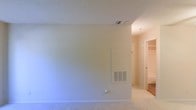 This single-floor residential space offers a clean, neutral aesthetic throughout. The interior features light-colored carpeting that runs through the main living area and into the connecting hallway, providing a cohesive and comfortable feel underfoot. Walls are finished in a soft, neutral tone, creating a bright and versatile backdrop suited to a variety of furnishing styles. The textured ceiling adds subtle dimension to the space, while ceiling-mounted HVAC vents and a wall-mounted ventilation unit indicate a well-integrated climate control system. A hallway visible from the main room leads to additional areas of the unit, including what appears to be a closeted storage or wardrobe space, suggesting practical organization options. The overall layout is straightforward and functional, with a simple, uncluttered design that allows the space to feel open and adaptable. The neutral color palette and carpeted flooring throughout contribute to a warm, understated ambiance, making this a space that is easy to personalize and move into with minimal preparation.