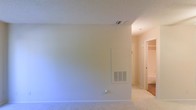 This single-floor residential space offers a clean, neutral aesthetic throughout. The interior features light-colored carpeting that runs through the main living area and into the connecting hallway, providing a cohesive and comfortable feel underfoot. Walls are finished in a soft, neutral tone, creating a bright and versatile backdrop suited to a variety of furnishing styles. The textured ceiling adds subtle dimension to the space, while ceiling-mounted HVAC vents and a wall-mounted ventilation unit indicate a well-integrated climate control system. A hallway visible from the main room leads to additional areas of the unit, including what appears to be a closeted storage or wardrobe space, suggesting practical organization options. The overall layout is straightforward and functional, with a simple, uncluttered design that allows the space to feel open and adaptable. The neutral color palette and carpeted flooring throughout contribute to a warm, understated ambiance, making this a space that is easy to personalize and move into with minimal preparation.