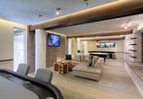 This single-floor space showcases a sophisticated, open-concept design that blends modern materials with thoughtful lighting. A striking textured stone accent wall serves as the focal point of the lounge area, anchoring a wall-mounted flat-screen television and providing rich visual contrast against the smooth, neutral-toned surfaces throughout. Structural columns with a curved, sculpted finish add architectural interest while subtly defining different zones within the open layout.

The lounge area is furnished with a generously sized sectional sofa in a warm beige tone, complemented by teal accent pillows and a distinctive rustic wood coffee table that introduces an organic element amid the contemporary design. Recessed track lighting and elegant cove lighting embedded in the ceiling create a layered, ambient atmosphere, casting a warm golden glow across the space.

Adjacent areas are partially visible, hinting at additional functional zones, including what appears to be a bar or counter area with pendant lighting. Large windows allow natural light to filter in, enhancing the open, airy feel. The overall ambiance is polished and inviting, suitable for both relaxation and social gatherings.