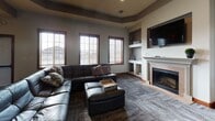 This single-floor space is thoughtfully designed to accommodate both social gatherings and everyday use, combining comfortable living, dining, and kitchen areas in one cohesive layout. The living room serves as a central gathering point, featuring a large sectional leather sofa, a tile-surround fireplace with built-in shelving on either side, and a wall-mounted television. Patterned carpet with a subtle herringbone texture runs underfoot, while multiple windows dressed with wood-framed horizontal blinds allow natural light to filter in and offer exterior views. Neutral gray walls and rich wood trim throughout create a warm, grounded aesthetic.

Two separate dining areas provide flexible seating for groups of varying sizes. Each is furnished with a substantial dark wood farmhouse-style table and matching chairs, set beneath tray ceilings with recessed lighting. Interior pass-through windows connect the spaces visually, enhancing an open and communicative flow between rooms.

The kitchen is well-equipped with warm cherry wood cabinetry, laminate countertops, tile flooring, and black appliances including a range, refrigerator with ice maker, and microwave. A central island with pendant lighting adds additional workspace. The overall design balances functionality with an inviting, residential character suitable for group gatherings.