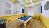 This energetic game room is designed to entertain, combining bold aesthetic choices with a well-rounded selection of recreational amenities. The space is immediately defined by its eye-catching color scheme — bright yellow wainscoting lines the lower walls, creating a lively contrast against the crisp white upper walls and ornate crown molding. Overhead, a coffered ceiling with yellow-painted recesses and a multi-shade chandelier adds architectural depth and a polished finishing touch.

The flooring features a geometric-patterned carpet in coordinating tones of brown and gold, reinforcing the room's dynamic visual identity. At the center of the space sits a white ornate billiard table with a navy felt surface, serving as the room's focal point. A foosball table provides additional entertainment, while a glass-top table with a built-in chessboard offers a quieter gaming option for smaller groups.

Wall-mounted blue acoustic panels complement a gallery of six framed colorful artworks, adding both functional sound absorption and decorative character. A wall-mounted flat-screen TV and a mounted cue rack further enhance the room's utility. Natural light filters in through a slatted window, and French doors provide a welcoming entry point into this thoughtfully curated recreational space.