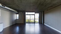 This single-floor space showcases a striking industrial aesthetic defined by its exposed concrete ceiling, which lends a raw, urban character to the interior. The ceiling's unfinished texture is complemented by visible structural columns and overhead ducting, reinforcing the space's contemporary loft-like character. Underfoot, rich dark hardwood flooring runs throughout, providing a warm contrast to the cooler tones above.

The main living area is bright and open, anchored by a generous wall of floor-to-ceiling sliding glass doors that draw in abundant natural light. These doors lead directly to a balcony enclosed by a sleek metal railing, offering views of the surrounding cityscape, including glimpses of neighboring buildings and mature trees below. A secondary window on the adjacent wall further enhances the natural light within the space.

The layout is open-plan, providing flexible use of the available area. The minimalist, unfurnished state of the space allows its architectural elements — the interplay between the raw concrete ceiling, the dark floors, and the expansive glazing — to take center stage, creating a versatile and modern environment.