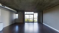 This single-floor space showcases a striking industrial aesthetic defined by its exposed concrete ceiling, which lends a raw, urban character to the interior. The ceiling's unfinished texture is complemented by visible structural columns and overhead ducting, reinforcing the space's contemporary loft-like character. Underfoot, rich dark hardwood flooring runs throughout, providing a warm contrast to the cooler tones above.

The main living area is bright and open, anchored by a generous wall of floor-to-ceiling sliding glass doors that draw in abundant natural light. These doors lead directly to a balcony enclosed by a sleek metal railing, offering views of the surrounding cityscape, including glimpses of neighboring buildings and mature trees below. A secondary window on the adjacent wall further enhances the natural light within the space.

The layout is open-plan, providing flexible use of the available area. The minimalist, unfurnished state of the space allows its architectural elements — the interplay between the raw concrete ceiling, the dark floors, and the expansive glazing — to take center stage, creating a versatile and modern environment.