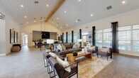 This well-appointed single-floor communal space combines modern farmhouse aesthetics with refined comfort across a thoughtfully designed open floor plan. The centerpiece of the interior is a dramatic vaulted ceiling adorned with white shiplap paneling and an exposed natural wood beam, lending warmth and architectural character to the expansive main lounge area. Recessed lighting and statement pendant fixtures illuminate the space, while large windows dressed with floor-length dark curtains and horizontal blinds invite generous natural light and views of the surrounding outdoors.

The main lounge is furnished with plush, dark-toned sofas and accent chairs arranged around a solid wood coffee table, all set atop a warm area rug — creating an inviting gathering space. Adjacent to the lounge, a dedicated seating area near the entry features neutral upholstered chairs and tiled flooring with a herringbone accent pattern. A built-in kitchenette area includes a large white shiplap island with a quartz-style countertop, white cabinetry, a mounted flat-screen television, and a patterned tile feature wall. A separate nook offers built-in white cabinetry with a dark countertop and floating wood shelves. The consistent use of wood-look tile flooring, neutral tones, and curated décor ties the entire space together seamlessly.