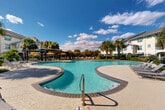 Located at 301 Lakeshore Blvd N in Slidell, Louisiana, this expansive outdoor amenity area showcases a generously sized freeform swimming pool as its centerpiece. The pool's curvilinear design creates a resort-like atmosphere, with crystal-clear turquoise water and a wide, stamped concrete deck that provides ample space for lounging and movement around the perimeter.

A prominent pergola structure positioned poolside offers shaded seating beneath yellow-accented umbrellas, making it an ideal gathering spot. Surrounding the pool, multiple chaise lounges are arranged throughout the deck and landscaped areas, accommodating a large number of guests comfortably.

The landscaping is lush and tropical in character, featuring tall palm trees, ornamental grasses, and manicured shrubs that create a sense of privacy and a distinctly Southern resort ambiance. The greenery provides a natural buffer, enhancing the overall aesthetic and atmosphere of the space.

The pool area is framed by multi-story residential buildings finished in warm yellow and blue tones, complete with private balconies that overlook the grounds. A decorative iron perimeter fence encloses the amenity space. Under the wide-open Louisiana sky, this outdoor area delivers a vibrant, sun-drenched setting well-suited for recreation and relaxation throughout the year.