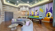 Located at 3500 Millenia Blvd in Orlando, Florida, this dynamic community lounge and game room delivers a bold, retro-inspired aesthetic that immediately captures attention. The single-level space features herringbone-patterned wood-look flooring that runs throughout, grounding the lively décor with a warm, cohesive foundation. Tray ceilings with teal accents and recessed lighting add architectural depth and a polished finish overhead.

A full-size pool table with a vivid blue felt surface serves as the centerpiece of the room, positioned beneath the layered ceiling detail. Flanking it is a curved, channel-tufted gray sectional sofa that invites relaxed socializing, complemented by a wall-mounted entertainment center with built-in shelving displaying colorful decorative objects and a flat-screen TV.

A bar counter with bar-height seating and warm accent lighting lines one wall, accented by horizontal wood slat paneling that adds texture and a casual, modern feel. The defining feature of the space is a sweeping, large-scale mural depicting stylized retro characters in vivid yellows, purples, and greens against a graphic patterned backdrop — giving the room an unmistakable, playful personality.

French doors with teal drapery allow natural light to filter in and provide access to the exterior. Colorful framed artwork and a tiki-style wall sculpture round out the eclectic, art-forward design of this energetic gathering space.