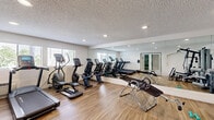 This fitness center in Escondido, California, offers a well-equipped exercise space designed to support a well-rounded workout routine. The single-room facility features warm hardwood-style flooring throughout, crisp white walls, and a textured ceiling with recessed lighting that keeps the space bright and functional. A large floor-to-ceiling mirror spans one wall, visually expanding the room and allowing users to monitor their form during exercise. Natural light filters in through multiple windows, contributing to an open and energizing atmosphere. Entry is through a set of elegant glass-paned French doors, adding a polished touch to the space. The cardio zone is well-appointed with multiple treadmills, an elliptical trainer, a stationary bike, and additional cardio machines, all positioned to take advantage of the natural light. The strength training area includes a multi-station cable machine, a rowing machine, and various other resistance equipment, providing options for a full-body workout. Wall-mounted accessories and a small waste station help maintain an organized environment. The overall ambiance is clean, functional, and inviting, making this fitness center a convenient and practical destination for daily exercise.