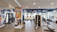 This contemporary fitness center is thoughtfully designed to accommodate a full spectrum of workout needs across a single, open floor. The space is divided into distinct functional zones, each outfitted with professional-grade equipment. The cardio area features a row of treadmills, elliptical machines, stair climbers, and stationary bikes, all positioned along large floor-to-ceiling windows that flood the space with natural light and offer expansive outdoor views. The strength training zone is equally well-appointed, housing a power rack with free weights, a comprehensive dumbbell rack, kettlebells, and an array of cable and resistance machines from leading equipment brands. Rubber flooring throughout provides both durability and comfort underfoot. The exposed ceiling, painted in a bold blue, reveals industrial-style ductwork and is fitted with modern pendant and linear lighting fixtures that give the space an urban, loft-like aesthetic. Striking abstract murals adorn several feature walls and structural columns, adding a vibrant artistic energy to the environment. A private studio room with frosted glass sliding doors offers space for focused training or stretching. The entry corridor is lined with a distinctive textured wall and includes water fountains for added convenience. Full-length mirrors amplify the sense of openness throughout.