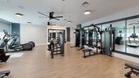 Located at 509 N Mangum St in Durham, North Carolina, this fitness center offers a comprehensive workout facility spread across a single floor. The space is divided into two distinct zones: a fully equipped gym area and a separate movement or group exercise studio.

The main gym area is generously sized and bathed in natural light from large windows fitted with roller shades. Light hardwood-style flooring runs throughout, creating a clean and energetic atmosphere. Large ceiling fans circulate air across the space, complementing the modern flush-mount lighting overhead. The room is outfitted with a wide array of strength machines, cable systems, free weight benches, treadmills, stair climbers, and other cardio equipment. Full-length mirrors along the walls enhance the sense of space and allow for proper form checks during workouts. Accent walls featuring geometric patterns add a contemporary design touch.

The adjacent movement studio provides an open, flexible area ideal for stretching, yoga, or functional training. It features the same light flooring and clean white walls, with a full-length mirror and stacked plyo boxes available for use. A large window allows natural light to fill the room. A corridor connects the two zones, lined with additional equipment and storage, ensuring a seamless flow throughout the facility.
