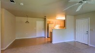 This single-floor residence offers a functional and welcoming layout centered around an open-concept living and kitchen area. The living space features soft, neutral-toned carpet underfoot, providing a comfortable setting that flows naturally toward the kitchen, where the flooring transitions to a smooth hard surface. A pendant light fixture adds a subtle decorative touch to the living area, while a ceiling fan enhances comfort overhead.

The kitchen is defined by warm, light wood cabinetry that extends along the upper walls, offering ample storage space. A peninsula-style counter separates the kitchen from the living area, creating a natural division while maintaining the open feel of the shared space. A full-size refrigerator and overhead lighting contribute to the kitchen's practical design.

The entryway is clearly defined by a solid white door, providing a clean and straightforward point of entry. Neutral-toned walls throughout the space keep the atmosphere bright and versatile, allowing the interior to feel open and adaptable. The overall layout is efficient and cohesive, making effective use of the available floor plan. The combination of soft carpeting in the living zone and hard flooring in the kitchen area adds both visual interest and practical utility to this well-organized single-level home.