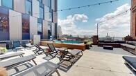 Located on Euclid Avenue in the heart of downtown Cleveland, this rooftop amenity level offers a well-rounded collection of indoor and outdoor spaces designed for relaxation and socializing. The centerpiece of the outdoor area is a wood-framed pool surrounded by sleek lounge chairs, ideal for soaking up the sun while taking in sweeping views of the Cleveland skyline. String lights overhead add a warm ambiance for evening gatherings. Adjacent to the pool, a cozy outdoor seating nook features wicker sectional sofas arranged around a fire pit table, set against an exposed brick wall that adds rustic character. A separate terrace area provides additional wicker seating with views of the surrounding urban architecture. The indoor lounge area is thoughtfully designed with a striking living green wall serving as the backdrop for a mounted flat-screen TV, flanked by rattan armchairs and a coffee table. A dedicated dining section offers multiple wooden tables with navy-cushioned chairs, accented by decorative wall art and potted tropical plants. The space is equipped with accessible features, including a lift alongside the exterior staircase, ensuring inclusivity throughout. Together, these amenities create a versatile, elevated retreat above the vibrant streets of Cleveland.