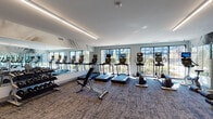 Located at 2100 Progress Pkwy in Schaumburg, Illinois, this contemporary fitness center is thoughtfully designed to support a full range of workout routines. The space is organized into distinct functional zones, offering both cardio and strength training areas across a single open floor.

The cardio zone is expansive, housing a generous lineup of treadmills and elliptical machines, each equipped with individual display screens for an engaging workout experience. Large, multi-pane windows line the perimeter walls, flooding the space with natural light and offering open views of the surrounding greenery — creating an energizing atmosphere for exercise.

The strength training area is equally well-appointed, featuring cable machines, a multi-functional cable crossover system, weight benches, a rowing machine, and a rack of dumbbells arranged along a mirrored wall. Full-length wall mirrors are positioned throughout the facility, visually expanding the space and allowing users to monitor their form during workouts.

Underfoot, durable textured carpet tiles provide comfort and traction throughout the floor. Linear LED lighting fixtures run along the ceiling, delivering consistent, bright illumination across all areas. The overall design aesthetic is clean and modern, with a neutral color palette of soft grays and whites that contributes to a focused and professional fitness environment.