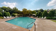 Located at 2900 Baby Ruth Ln in Nashville, Tennessee, this outdoor amenity area centers around a generously sized swimming pool with clear, blue-green water. The pool is finished with a broad concrete surround that provides ample space for lounging, lined with rows of reclining chaise lounges and patio chairs arranged for both relaxation and socializing. A shaded seating area with a large umbrella and table set offers a comfortable retreat from the sun.

The pool area is anchored by a classic brick clubhouse building featuring a pitched tile roof, large windows, and a welcoming facade that reflects a traditional residential architectural style. A raised deck with steps connects the building to the pool deck, and a wooden pergola structure adds architectural interest to one end of the space.

The entire area is enclosed by a secure perimeter fence and framed by mature trees, manicured shrubs, and well-maintained landscaping that provides privacy and a lush, green backdrop. Ornamental globe lampposts are positioned throughout the deck for evening illumination. Together, these elements create a well-appointed, resort-style outdoor gathering space suited for both active recreation and relaxed outdoor leisure.