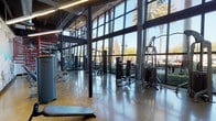 This fitness center is a thoughtfully designed, single-floor facility that combines industrial aesthetics with an open, energizing atmosphere. The most striking architectural feature is the expansive floor-to-ceiling glass wall that lines one side of the space, flooding the interior with abundant natural light and offering panoramic views of mature palm trees and lush outdoor landscaping. The soaring ceiling height amplifies the sense of openness, with exposed ductwork and structural steel beams contributing to the space's bold industrial character. A dramatic red spiral staircase serves as a visual centerpiece, leading to an upper mezzanine level visible from the main floor. Polished concrete floors run throughout, reflecting light and adding to the clean, contemporary feel. The strength training area is comprehensively outfitted with a wide selection of cable and resistance machines positioned along the windowed wall, while a dedicated free weights zone features adjustable benches, dumbbells, and barbells set on protective rubber matting. Vibrant large-scale murals depicting athletic figures accent the interior walls, adding a dynamic and motivational visual element. The facility is well-lit with modern linear lighting, and the layout provides ample space between equipment for comfortable, unrestricted movement throughout the workout floor.
