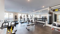 Located at 589 Bedford St in Stamford, Connecticut, this fitness center offers a well-appointed, single-floor exercise space designed to support a comprehensive workout experience. The room features durable rubber flooring throughout, providing a safe and functional surface for a wide range of activities.

The cardio section is equipped with multiple treadmills and elliptical machines, each featuring integrated display screens for performance tracking. Large windows along the walls allow ample natural light to fill the space, creating an energizing atmosphere for workouts. Recessed ceiling lighting supplements the natural light for consistent illumination throughout.

The strength training area includes a full dumbbell rack with a wide range of weights, a flat weight bench, and a cable/multi-use strength machine. A large wall-mounted mirror runs the length of the strength zone, enhancing the sense of space and allowing users to monitor their form. A heavy punching bag mounted on a wall-mounted rack adds a boxing and functional training element to the facility.

Additional equipment includes stability balls and resistance accessories. A glass-door entry provides access to the space, with a utility enclosure tucked neatly into one corner. The overall design is clean and modern, with white walls and a neutral color palette that keeps the environment focused and professional.