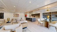 Located at 1301 Burnside Rd W in View Royal, BC, this thoughtfully designed single-floor amenity space combines a fully equipped kitchen, a generous living area, and a dining zone in one seamless open-concept layout. Light hardwood flooring runs throughout, complementing the bright, neutral palette and recessed ceiling lighting that illuminates the entire space.

The kitchen is a standout feature, showcasing two-tone cabinetry in light wood and charcoal, white quartz countertops, a mirrored backsplash, and gold-accented ring hardware. A large central island with integrated seating provides ample space for gathering, while a dramatic cluster of pendant lights overhead adds a refined, modern touch. Full-size appliances, including a refrigerator and built-in microwave, complete the functional setup.

The living area is anchored by a sculptural textured feature wall housing a sleek linear fireplace below a wall-mounted television, flanked by vertical wood slat panels and elegant wall sconces. Plush cream and caramel-toned seating arrangements, accented with marble side tables and gold décor, create a warm and inviting atmosphere. Large windows along one wall invite natural light and offer views of the surrounding greenery. A dedicated dining area with a glass-top table and white upholstered chairs rounds out this sophisticated and versatile communal space.
