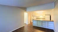 This single-floor residence offers a thoughtfully designed open-concept layout that connects the living area and kitchen in a cohesive, functional flow. The kitchen is a standout feature, showcasing white cabinetry that extends to the upper walls, a dark-toned countertop peninsula with built-in open shelving beneath, and white appliances including a range and microwave. A pendant light fixture illuminates the workspace, complementing the bright, clean aesthetic of the room. The kitchen also includes a sink positioned along the back counter, making it both practical and well-appointed. Dark hardwood flooring runs throughout the space, providing a rich contrast against the soft, neutral gray walls and crisp white trim and baseboards. The open living area offers generous room to arrange furnishings, benefiting from the seamless sight lines into the kitchen. A hallway leads to additional rooms, suggesting a well-organized floor plan. The overall ambiance is bright, clean, and move-in ready, with a neutral palette that provides a versatile backdrop for a variety of interior styles. The space balances modern touches with approachable, comfortable design throughout.