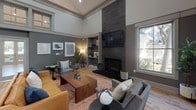 Located at 4606 Cedar Springs Rd in Dallas, TX, this single-floor space offers a thoughtfully designed interior that blends contemporary style with warm, inviting details. The layout is anchored by a generously proportioned lounge area defined by soaring ceilings and a dramatic floor-to-ceiling stone fireplace, which serves as the room's visual centerpiece. A wall-mounted television above the fireplace and built-in shelving niches with accent lighting add both function and character to the space. The lounge is furnished with a mix of leather and upholstered seating arranged around a solid wood coffee table, all set atop a rich, patterned area rug over light hardwood-style flooring that runs throughout. Natural light filters in through multiple windows, offering views of the surrounding greenery. Adjacent to the lounge, a round dining table with modern chairs provides a dedicated area for gathering. The kitchen features dark cabinetry, stainless steel appliances, and a large island with a white countertop and decorative paneling, flanked by sleek bar-height seating — ideal for casual interaction. Neutral gray walls throughout create a cohesive, sophisticated palette, complemented by curated artwork. The overall ambiance is modern yet comfortable, well-suited for community use.