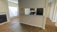 This single-floor residence offers a functional and well-designed open-concept layout that seamlessly connects the kitchen and living areas. The kitchen is equipped with a full suite of appliances, including a black refrigerator, a gas range with a stainless steel microwave above, and ample cabinet storage finished in a light tone. A raised breakfast bar with granite-style countertops serves as a natural divider between the kitchen and the adjacent living space, providing both a casual dining option and a visual anchor for the open floor plan. The living area is finished with warm, wood-style flooring that adds character and cohesion throughout the space. Natural light filters into the living area through a window fitted with blinds, brightening the neutral-toned walls and enhancing the overall sense of openness. White baseboards provide a clean contrast to the flooring and walls, contributing to a polished, well-maintained aesthetic. The layout is practical and efficient, making excellent use of the available space while maintaining a comfortable and inviting atmosphere suitable for everyday living.