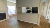 This single-floor residence offers a functional and well-designed open-concept layout that seamlessly connects the kitchen and living areas. The kitchen is equipped with a full suite of appliances, including a black refrigerator, a gas range with a stainless steel microwave above, and ample cabinet storage finished in a light tone. A raised breakfast bar with granite-style countertops serves as a natural divider between the kitchen and the adjacent living space, providing both a casual dining option and a visual anchor for the open floor plan. The living area is finished with warm, wood-style flooring that adds character and cohesion throughout the space. Natural light filters into the living area through a window fitted with blinds, brightening the neutral-toned walls and enhancing the overall sense of openness. White baseboards provide a clean contrast to the flooring and walls, contributing to a polished, well-maintained aesthetic. The layout is practical and efficient, making excellent use of the available space while maintaining a comfortable and inviting atmosphere suitable for everyday living.