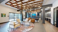 Located at 2870 Pearson Pkwy in Plainfield, Indiana, this generously sized single-floor amenity space offers a sophisticated and modern environment designed for socializing and recreation. The interior is defined by a striking coffered ceiling with exposed wooden beams and an abundance of Edison-style pendant lights, creating a warm, inviting ambiance throughout.

The main lounge area features multiple seating arrangements, including rich cognac leather sofas and armchairs paired with contemporary accent chairs, layered over colorful area rugs atop large-format tile flooring. A dramatic mosaic-tiled double-sided fireplace serves as a central focal point, flanked by mounted flat-screen televisions. Large floor-to-ceiling windows and sliding glass doors flood the space with natural light and provide direct access to an outdoor patio equipped with grilling stations.

A dedicated bar area showcases a long marble-topped counter with leather barstools, stylish pendant lighting, and colorful artwork. Adjacent spaces include a billiards room with a full-sized pool table bathed in natural light, a shuffleboard table, and an arcade game station. Textured accent walls in iridescent tile add depth and visual interest throughout. The overall design seamlessly blends industrial and contemporary elements for a dynamic, community-centered atmosphere.