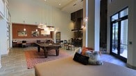 This single-floor amenity lounge is designed to accommodate a variety of social activities within one expansive, open-plan space. The room immediately impresses with its soaring ceilings, which lend an airy, grand atmosphere throughout. A full-sized billiards table anchors the central game area, positioned atop a bold geometric patterned area rug and illuminated by a cluster of rectangular pendant lights that add a warm, layered glow.

Adjacent to the game zone, a comfortable lounge seating area features plush cushions and textured throw pillows in rust and neutral tones, creating an inviting space to relax. Moving deeper into the room, a bar and dining area offers high-top seating along a sleek counter, framed by tall dark cabinetry and accented with globe pendant lights that introduce a modern, industrial touch. Exposed brick detailing adds warmth and texture to this section of the space.

The overall design palette blends warm terracotta and earthy tones with dark wood accents and light-toned walls, resulting in a sophisticated yet approachable ambiance well-suited for both casual socializing and structured entertainment.