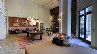 This single-floor amenity lounge is designed to accommodate a variety of social activities within one expansive, open-plan space. The room immediately impresses with its soaring ceilings, which lend an airy, grand atmosphere throughout. A full-sized billiards table anchors the central game area, positioned atop a bold geometric patterned area rug and illuminated by a cluster of rectangular pendant lights that add a warm, layered glow.

Adjacent to the game zone, a comfortable lounge seating area features plush cushions and textured throw pillows in rust and neutral tones, creating an inviting space to relax. Moving deeper into the room, a bar and dining area offers high-top seating along a sleek counter, framed by tall dark cabinetry and accented with globe pendant lights that introduce a modern, industrial touch. Exposed brick detailing adds warmth and texture to this section of the space.

The overall design palette blends warm terracotta and earthy tones with dark wood accents and light-toned walls, resulting in a sophisticated yet approachable ambiance well-suited for both casual socializing and structured entertainment.