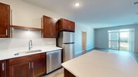 Located at 1875 Paulson Rd in River Falls, Wisconsin, this two-bedroom, one-bathroom residence offers a well-designed single-floor layout with a thoughtful blend of modern finishes and functional living spaces. The open-concept kitchen and living area forms the heart of the home, anchored by a striking kitchen featuring rich, dark-stained wood cabinetry, white countertops, a central island ideal for dining or meal prep, and a full suite of stainless steel appliances including a refrigerator, dishwasher, range, and over-the-range microwave. Light wood-tone flooring runs throughout the main living areas, contributing to a clean, contemporary aesthetic. The spacious living room benefits from large sliding glass doors that open onto a private balcony, flooding the space with natural light and offering pleasant outdoor views. Both bedrooms are carpeted for comfort, with the primary bedroom providing a generous footprint and convenient closet access. The full bathroom is well-appointed with dark wood vanity cabinetry, white countertops, a tub-shower combination, and light wood-tone flooring. Neutral wall tones throughout create a calm and versatile backdrop, making this a well-rounded and move-in-ready residence.