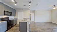 This single-floor residence offers a thoughtfully designed open-concept layout that seamlessly connects the kitchen and living areas. The kitchen serves as the heart of the home, showcasing gray shaker-style cabinetry, light granite countertops, and a matching granite-topped center island with a built-in sink. Black stainless appliances — including a refrigerator, electric range, and over-the-range microwave — complete the kitchen's contemporary aesthetic. Track lighting overhead adds both function and style to the space. Durable hardwood-style flooring runs throughout the main living and kitchen areas, providing a clean, modern foundation. The living area is bright and inviting, featuring a ceiling fan with integrated lighting and a door leading to an outdoor area, allowing natural light to fill the room. The bedrooms are finished with plush carpeting and ceiling fans, offering a comfortable and quiet retreat. Each bedroom benefits from window access, bringing in additional natural light. The bathroom includes a full-size soaking tub and shower combination with clean white surroundings and tile flooring. Neutral warm-toned walls throughout the residence create a cohesive and welcoming ambiance, ready to accommodate a variety of interior styles.