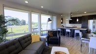 This single-floor space showcases a thoughtfully designed open-concept layout that seamlessly connects the living area and kitchen. The living space is anchored by large, floor-to-ceiling windows that flood the interior with abundant natural light while framing sweeping views of a lush green landscape and what appears to be a marina in the distance. A sliding glass door provides direct access to an outdoor balcony, further integrating the interior with the scenic surroundings.

The living area features contemporary furnishings, including a tufted gray sofa and a matching armchair, complemented by a statement arc floor lamp. Light hardwood flooring extends throughout, adding warmth to the neutral color palette.

The kitchen is finished in a sleek, modern style with dark cabinetry, a gas cooktop on a central island, and a stainless steel range hood overhead. Pendant lighting illuminates the island, which is lined with white bar stools for casual dining. The overall ambiance of the space is clean, contemporary, and bright, making excellent use of its elevated position to maximize natural light and outdoor views.