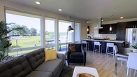 This single-floor space showcases a thoughtfully designed open-concept layout that seamlessly connects the living area and kitchen. The living space is anchored by large, floor-to-ceiling windows that flood the interior with abundant natural light while framing sweeping views of a lush green landscape and what appears to be a marina in the distance. A sliding glass door provides direct access to an outdoor balcony, further integrating the interior with the scenic surroundings.

The living area features contemporary furnishings, including a tufted gray sofa and a matching armchair, complemented by a statement arc floor lamp. Light hardwood flooring extends throughout, adding warmth to the neutral color palette.

The kitchen is finished in a sleek, modern style with dark cabinetry, a gas cooktop on a central island, and a stainless steel range hood overhead. Pendant lighting illuminates the island, which is lined with white bar stools for casual dining. The overall ambiance of the space is clean, contemporary, and bright, making excellent use of its elevated position to maximize natural light and outdoor views.