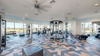 Located at 1902 Millenia Ave in Chula Vista, California, this well-appointed fitness center offers a bright, open, and thoroughly equipped workout environment spread across a single floor. The space is defined by its striking chevron-patterned carpet in shades of blue, gray, and tan, which adds a modern, energetic aesthetic throughout.

Large floor-to-ceiling windows line the perimeter, inviting generous natural light and offering views of the surrounding palm tree-dotted landscape. Ceiling fans and recessed lighting complement the airy atmosphere, keeping the space comfortable during any workout.

The fitness floor is comprehensively outfitted with a wide variety of Life Fitness equipment, including treadmills, ellipticals, stationary bikes, stair climbers, and rowing machines for cardio training. A dedicated strength zone features a multi-functional cable machine, free weights, kettlebells, resistance equipment, and weight benches to support full-body training routines.

Floor-to-ceiling mirrors are strategically placed along multiple walls, enhancing the sense of spaciousness while allowing users to monitor their form. A designated training zone includes medicine balls, stability equipment, and additional functional fitness tools. A large flat-screen television is mounted prominently for entertainment during workouts. The clean white walls and modern architectural details round out this polished, high-functioning fitness facility.