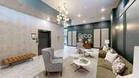 The Deco, located at 1301 Mulberry St in Des Moines, Iowa, is a thoughtfully designed residential building whose common areas exude refined elegance and modern sophistication. The grand entrance lobby immediately sets the tone, featuring polished marble-look tile flooring, soaring ceilings, and a striking sculptural chandelier with gold-toned arms and globe lights. Deep teal-painted wainscoting with decorative paneling lines the walls, complemented by warm brass wall sconces that cast an inviting glow. The lounge area is furnished with tufted olive-green sofas, patterned accent chairs, and sleek marble-top coffee tables on gold bases, all anchored by a geometric patterned area rug — creating a comfortable yet upscale gathering space. A dedicated mailroom, accessible from the entrance corridor, features floor-to-ceiling glass wall panels with black steel frames that flood the space with natural light, alongside a full bank of resident mailboxes. The fitness center is well-equipped with cardio machines, a rowing machine, cable weight system, free weights, and flat-screen monitors, all set on durable rubber flooring with high ceilings and exterior-facing windows. The building also includes a balcony, extending the living experience outdoors. Throughout, the design seamlessly combines Art Deco inspiration with contemporary finishes.