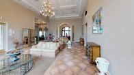 This sophisticated lobby exudes refined elegance through its carefully curated architectural details and classic décor. Soaring ceilings with coffered tray detailing and crown molding establish a grand first impression, while a stunning multi-arm chandelier serves as a luminous centerpiece overhead. The expansive space is grounded by large-format terracotta tile flooring arranged in a classic diamond pattern, adding warmth and texture throughout.

A striking ornate white fireplace with intricate carved detailing and a gilded mirror above serves as a focal point in the seating area, complemented by comfortable wingback chairs and upholstered sofas arranged to encourage conversation. Decorative wall art and sculptural relief panels add layers of visual interest to the neutral, warm-toned walls.

Generous windows dressed with rich, draped swag curtains flood the lobby with natural light and offer views of the surrounding outdoor pool area and lush landscaping. A graceful arched entryway leads deeper into the facility, while a glass-paned French door provides access to an adjacent game or meeting room. The overall ambiance is welcoming and upscale, blending classical architectural elements with comfortable furnishings to create a distinguished and inviting communal space.