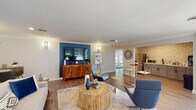 Located at 1331 Wicklow Ct in Cary, NC, this single-floor space offers a thoughtfully designed open-concept layout that seamlessly connects multiple functional areas. The main living area is anchored by a striking deep blue accent wall, which serves as a bold focal point against the neutral gray walls and white crown molding throughout. Wood-look flooring runs continuously across the space, contributing to a cohesive and warm aesthetic.

The living area is generously sized, accommodating multiple seating arrangements centered around a distinctive cylindrical wood coffee table atop a layered area rug. Wall-mounted sconces provide ambient lighting on either side of the accent wall, complementing the recessed ceiling lights above. A wooden credenza beneath the mounted flat-screen television adds functional storage and visual warmth.

Adjacent to the living area, a compact kitchenette features gray cabinetry with matte black hardware, a white countertop, a stainless steel refrigerator, and a microwave. The backsplash consists of a geometric 3D tile pattern in neutral tones, adding texture and visual interest to the space.

The dining area is positioned near a wall of large sliding glass doors and expansive windows dressed in blue floral curtains, flooding the space with natural light and offering direct views of the landscaped outdoor surroundings.