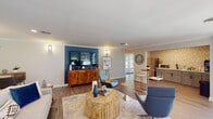 Located at 1331 Wicklow Ct in Cary, NC, this single-floor space offers a thoughtfully designed open-concept layout that seamlessly connects multiple functional areas. The main living area is anchored by a striking deep blue accent wall, which serves as a bold focal point against the neutral gray walls and white crown molding throughout. Wood-look flooring runs continuously across the space, contributing to a cohesive and warm aesthetic.

The living area is generously sized, accommodating multiple seating arrangements centered around a distinctive cylindrical wood coffee table atop a layered area rug. Wall-mounted sconces provide ambient lighting on either side of the accent wall, complementing the recessed ceiling lights above. A wooden credenza beneath the mounted flat-screen television adds functional storage and visual warmth.

Adjacent to the living area, a compact kitchenette features gray cabinetry with matte black hardware, a white countertop, a stainless steel refrigerator, and a microwave. The backsplash consists of a geometric 3D tile pattern in neutral tones, adding texture and visual interest to the space.

The dining area is positioned near a wall of large sliding glass doors and expansive windows dressed in blue floral curtains, flooding the space with natural light and offering direct views of the landscaped outdoor surroundings.