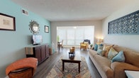 Located at 2545 NE Coachman Rd in Clearwater, Florida, this single-floor residence offers a well-organized and comfortable layout with a cohesive coastal-inspired design throughout. The living room serves as the heart of the home, featuring rich wood-style flooring, a textured ceiling, and a large picture window fitted with blinds that invites generous natural light. A teal accent wall adds a pop of color, complementing the warm, neutral tones found throughout the space. The living area flows seamlessly into the dining room, which opens directly to the kitchen via a pass-through counter, creating an connected, open feel ideal for entertaining. The kitchen is equipped with white cabinetry, dark countertops, a stainless steel sink, a tile mosaic backsplash, and a full suite of appliances. Both bedrooms are carpeted and feature louvered bi-fold closets and windows that provide natural light. The primary bedroom includes a private en-suite bathroom with a vanity and tub-shower combination. A second full bathroom with a tub-shower is also present. Wood-style flooring extends through the main living areas, tying the space together with a clean, modern aesthetic throughout this well-appointed Clearwater residence.