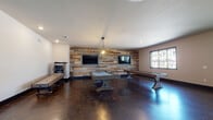 This single-story community clubhouse offers a well-rounded recreational and social environment, thoughtfully designed with a mix of modern and rustic elements throughout. The centerpiece of the space is a spacious game room featuring polished concrete floors, a striking reclaimed wood accent wall, and dual wall-mounted flat-screen TVs. A pool table and shuffleboard table anchor the room, while a vintage arcade machine and cue rack add to the entertainment offerings. A pendant light fixture provides warm, focused illumination over the game area, complementing the natural light that filters through large windows. Adjacent to the game room is a comfortable lounge area furnished with plush gray sofas and armchairs arranged around a coffee table, topped by a large ceiling fan and accented by contemporary wall art. A separate ping-pong room features dual ceiling fans, a teal accent wall with decorative metalwork, and ample open floor space. The clubhouse also includes a casual dining nook with bar-height tables and stools near glass doors that open to an outdoor area, as well as a fully equipped kitchen with dark cabinetry, patterned tile backsplash, stainless steel appliances, and direct access to an outdoor balcony with open views.