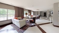 This expansive single-floor space is thoughtfully designed to accommodate both relaxed socializing and casual dining. The open layout is unified by a striking white floor accented with bold dark brown geometric inlay lines, creating a polished, contemporary aesthetic throughout.

The lounge area is anchored by tufted cream sofas and dark leather accent chairs arranged around a wooden coffee table atop a vibrant multicolored area rug. Tall floor lamps with warm gold shades flank large windows dressed in layered taupe drapes, allowing generous natural light to filter in while offering views of surrounding greenery. A sculptural gold starburst ceiling fixture adds an elegant focal point overhead.

The dining area transitions seamlessly from the lounge and features multiple white tables paired with white cane-back chairs, providing ample seating for gatherings. A display ledge along one wall showcases framed artwork, adding a curated touch. Black-framed glass doors open directly to an outdoor patio, connecting the interior to an exterior seating area and enhancing the sense of openness. The neutral, light-toned walls and high ceilings throughout contribute to an airy, welcoming ambiance well-suited for community events and everyday use.
