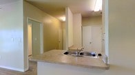 This single-floor residential space offers a practical and well-connected layout designed for comfortable everyday living. The kitchen serves as a central hub, featuring a peninsula countertop with a stainless steel sink that opens directly to the living area, creating an open-concept flow ideal for both daily routines and casual entertaining. White cabinetry lines the kitchen, providing ample storage, while the light-toned countertops add a clean, neutral aesthetic throughout. The open sightlines from the kitchen extend into the adjacent living and dining areas, enhancing the sense of space and connectivity. Warm, neutral-toned walls run throughout the unit, contributing to a calm and cohesive ambiance. Multiple doorways visible from the main living area lead to additional rooms, including what appears to be a bedroom and closet spaces, suggesting a thoughtful and efficient floor plan. The space is illuminated by ceiling-mounted light fixtures that provide consistent interior lighting. The flooring transitions between carpeted areas in the living spaces and other surfaces near the kitchen, adding texture and definition to each zone. Overall, this unit presents a straightforward and functional residential layout suited for a variety of lifestyles.