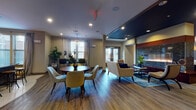 This single-floor amenity space is thoughtfully designed to offer a variety of social and recreational environments within one cohesive layout. The main lounge area features warm hardwood-style flooring, recessed lighting, and a dramatic floor-to-ceiling stone-clad fireplace that serves as a striking focal point. Adjacent to the fireplace is a relaxed seating arrangement with plush armchairs, a rustic wooden coffee table, and a built-in bar-height counter with industrial stools — ideal for casual gatherings. A decorative wall panel adds an artistic touch to this area. The open dining section accommodates multiple seating configurations, including a round table with mid-century modern chairs and a bar-height glass table, all bathed in natural light from large windows dressed with flowing linen curtains. A dedicated private screening room offers tiered leather recliner seating, a large wall-mounted display, teal drape accents, and soft wall sconces for an immersive cinematic experience. The fully equipped catering kitchen features dark cabinetry, rich wood countertops, a mosaic tile backsplash, beverage coolers, a large island with pendant lighting, and light tile flooring. A separate private dining or meeting room rounds out the space, complete with a glass-top table, a bold chandelier, and generous window views.