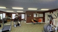 This single-floor fitness and wellness space is designed to provide a functional environment for physical activity and health-focused activities. The room features bold, dark-red walls that create a defined and energetic atmosphere, complemented by a textured ceiling with fluorescent lighting that ensures the space is well-lit throughout.

The fitness area is equipped with a cardio machine — a True-brand elliptical or stepper — positioned near the windows, which are fitted with slatted blinds that allow for controlled natural light. The patterned carpet, featuring an earthy floral design, covers the floor throughout, adding a layer of comfort underfoot.

A dedicated consultation or rest area sits along one wall, furnished with a small table and two orange chairs, creating a space for discussion, review, or brief rest between activities. Nearby, anatomical reference posters are displayed on the wall, reinforcing the health and wellness purpose of the room. A wall-mounted device — likely a thermostat or hand sanitizer dispenser — is also visible, reflecting practical amenities built into the space.

A wall-mounted clock and additional informational boards round out the functional details of this well-organized, purpose-driven fitness room, making it a comprehensive space for supporting an active and health-conscious lifestyle.