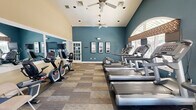 This fitness center offers a comprehensive workout environment designed to accommodate a variety of exercise routines. The space is characterized by soaring vaulted ceilings painted in a crisp cream tone, contrasted by bold teal-blue accent walls that create an energizing atmosphere. Ceiling fans are positioned throughout to maintain airflow and comfort during workouts.

The main floor is covered in multi-toned carpet tiles, providing a durable and functional surface. A row of treadmills equipped with individual monitors lines one side of the room, facing large arched and rectangular windows that allow generous natural light and outdoor views. Stationary bikes and additional cardio equipment are arranged on the opposite side, complemented by a wall-mounted mirror that visually expands the space.

A dedicated free weights area features a full rack of dumbbells in varying weights, a stability ball, and a flat workout bench, catering to strength training needs. A stretching and floor exercise zone is also present, equipped with folding chairs and exercise mats.

An interior window cutout connects two sections of the fitness center, enhancing the sense of openness. The overall design balances functionality with a welcoming aesthetic, making it a practical and motivating space for daily fitness activity.